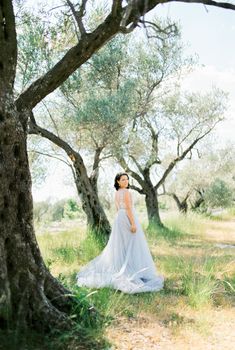Bride in a blue dress stands half-turned on a green meadow near the trees. High quality photo