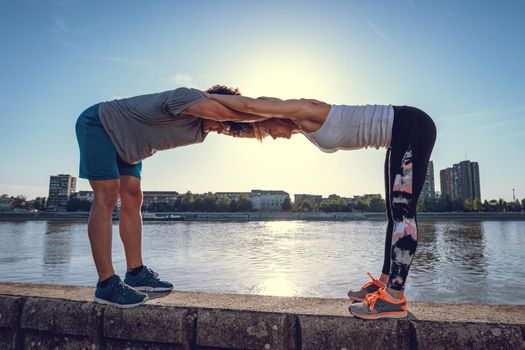 Young happy couple training outdoors by the river, stretching together on the wall at sunset.