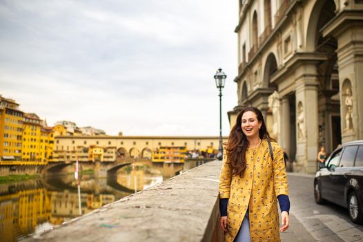 A tourist girl with an orange coat happily walks on the embankment in Florence, Italy.