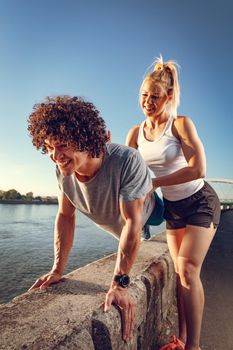 Young fitness couple doing workout on the wall  by the river in a sunset. The man is crouching and holding kettlebell, and the woman support him.