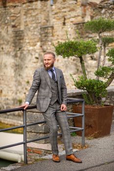 A man with a beard in a strict grey three-piece suit with a tie in the old town of Sirmione, a Stylish man in a grey suit in Italy.