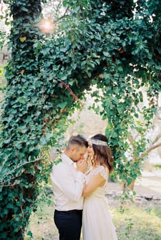 Groom kisses the hands of bride standing under an ivy-covered tree in the park. High quality photo
