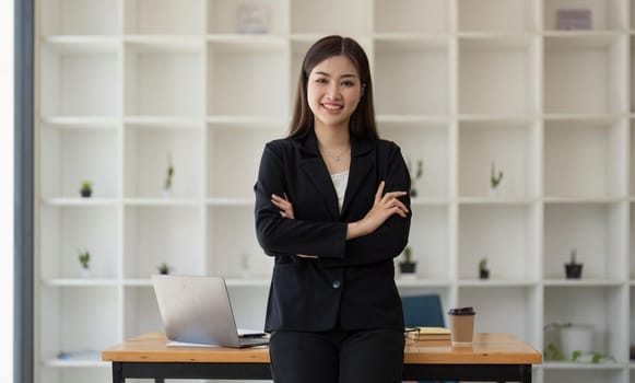 Young successful woman entrepreneur or an office worker stands with crossed arms near a desk in a modern office, looking at the camera and smiling.