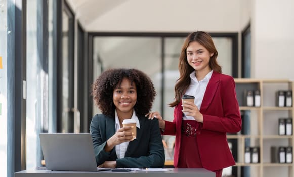 Two Asian business women enjoy chatting during coffee breaks at their desks in a comfortable conference room.