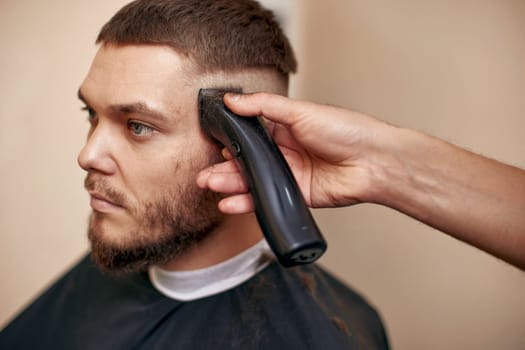 Barber trim hair with clipper on handsome bearded man in barber shop.