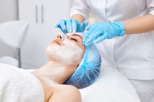 beautician applies the mask to the face of beautiful young woman in the spa salon. facial skin care concept.