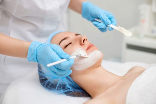 beautician applies the mask to the face of beautiful young woman in the spa salon. Anti-aging, facial skin care concept.