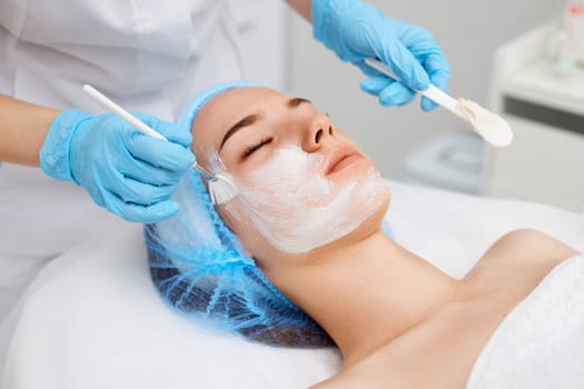 beautician applies the mask to the face of beautiful young woman in the spa salon. Anti-aging, facial skin care concept.