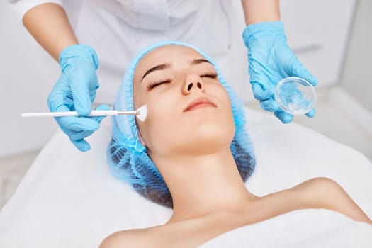 beautician applies the mask to the face of beautiful young woman in the spa salon. Anti-aging, facial skin care concept.