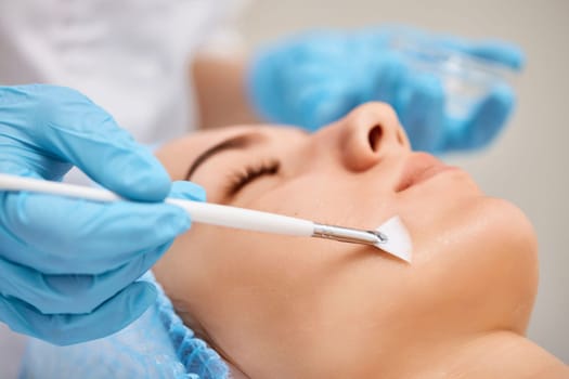 beautician applies the mask to the face of beautiful young woman in the spa salon. Anti-aging, facial skin care concept.