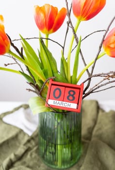 A very beautiful spring bouquet in a green vase stands on a table on a linen tablecloth, orange peony tulips. Wooden cubes with the inscription March 8