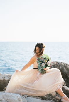 Bride with hair fluttering in the wind adjusts her dress sitting on the rocks by the sea. High quality photo