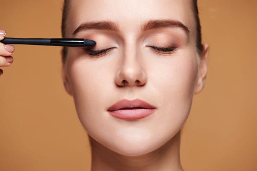young woman applying eyeshadows on the eyelids with make-up brush on beige background.