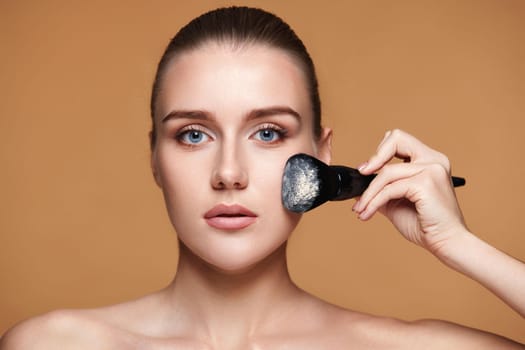 pretty young woman applying powder with professional make-up brush on beige background
