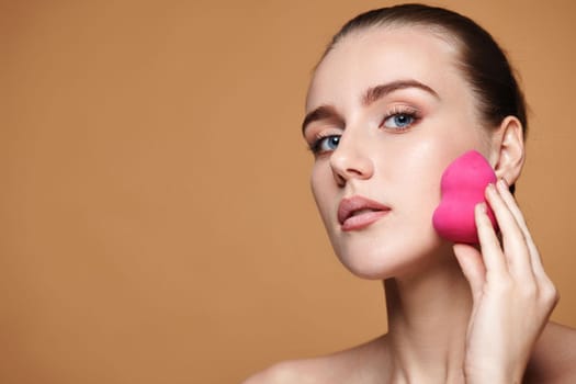 attractive young woman using pink cosmetic sponges on beige background. copy space