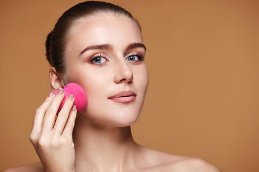smiling woman applying foundation on skin with beauty blender on beige background