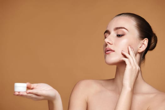 charming woman applying skin care cream on her face and holding jar of cosmetic product on beige background. copy space
