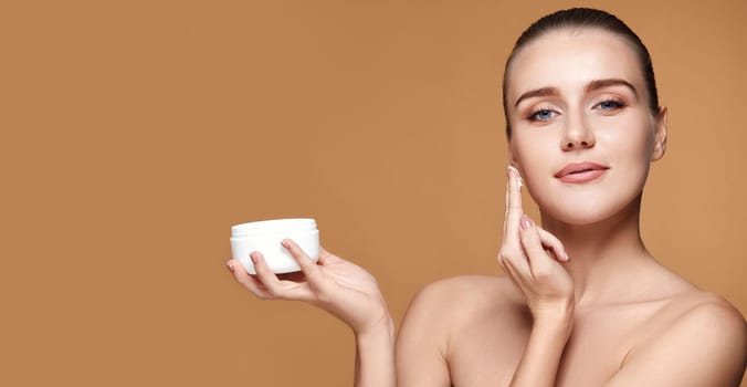 beautiful woman applying skin care cream on her face and holding jar of cosmetic product on beige background. copy space