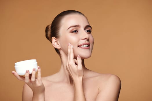 beautiful smiling young woman applying skin care cream on her face on beige background