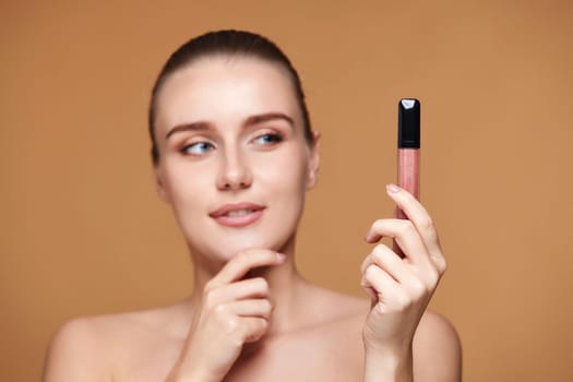 thinking smiling woman look away at nude lip gloss isolated on beige pastel background. focus on lip gloss