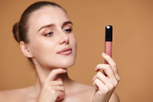 beautiful thinking woman look away at nude lip gloss isolated on beige pastel background. focus on lip gloss