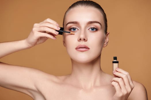 portrait of beautiful woman applying concealer under her eyes on beige background