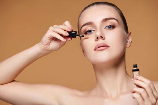 portrait of beautiful woman applying concealer under her eyes on beige background