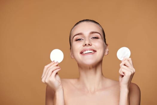 very excited laughing young woman holding two white cotton disks pad with micellar water on beige background