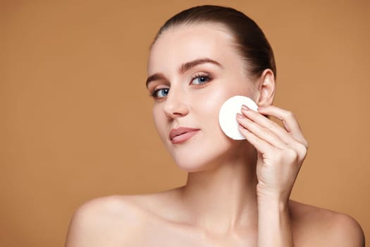 young woman holding white cotton pad with micellar water on beige background