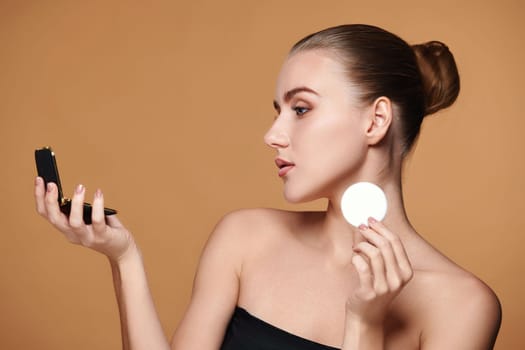 beautiful woman look at mirror and holding white cotton pad for removing makeup on beige background