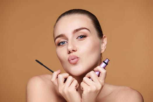 attractive young woman holding mascara and sending air kiss on beige background