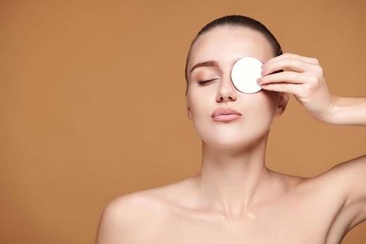 beautiful woman with naked shoulders close one eye with white cotton pad on beige background