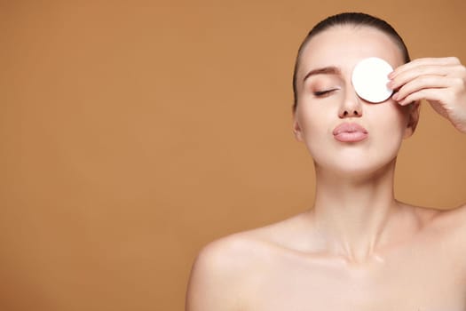 beautiful woman with naked shoulders close one eye with white cotton pad on beige background