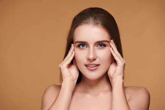 Beautiful woman with long hair and perfect clean skin touching face on beige background