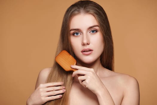 Beautiful brunette woman looks at the camera and combs her long natural hair with wooden brush on beige background