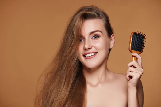 Beautiful hapy brunette woman showing wooden brush for healthy long hair on beige background