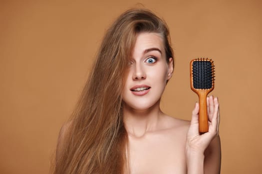 surprised beautiful brunette woman showing wooden brush for healthy long hair on beige background