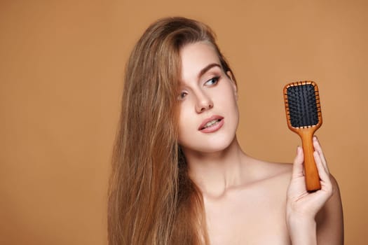 Beautiful brunette woman showing wooden brush and recommend haircare product for healthy long hair on beige background