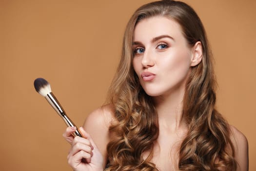 beautiful young curly woman holding make-up brush with powder on beige background