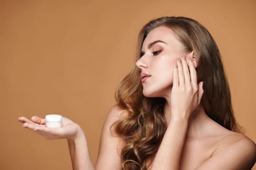 beautiful young woman applying skin care cream on her face on beige background