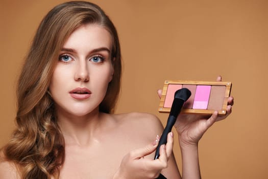 young woman applying blush on cheekbone with make-up brush and holding makeup palette on beige background. contouring