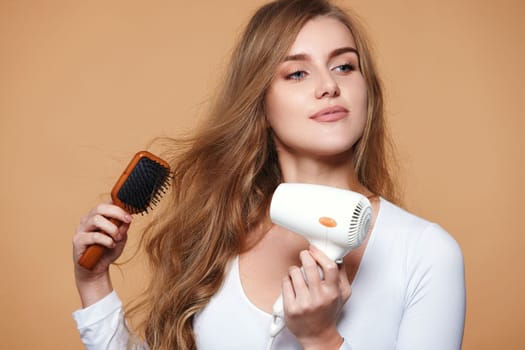 beautiful young happy woman drying strong healthy curly hair with dryer on beige background