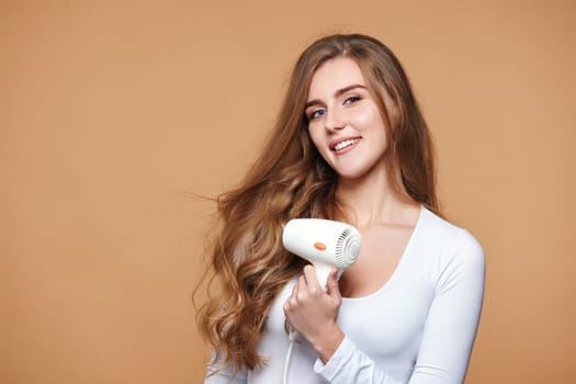 lovely woman with strong healthy curly hair using white hair dryer on beige background