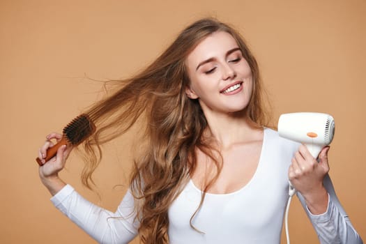 heerful girl with strong healthy curly hair using white hair dryer on beige background