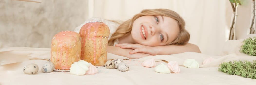 A girl with long hair in a light dress is sitting at the Easter table with cakes, spring flowers and quail eggs. Happy Easter celebration