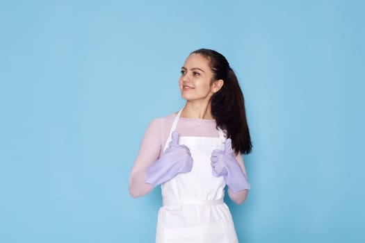 beautiful caucasian woman in violet rubber gloves and cleaner apron showing ok gesture on blue background. cleaning