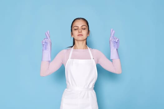 beautiful caucasian woman in rubber gloves with finger crossed gesture on blue background. cleaning