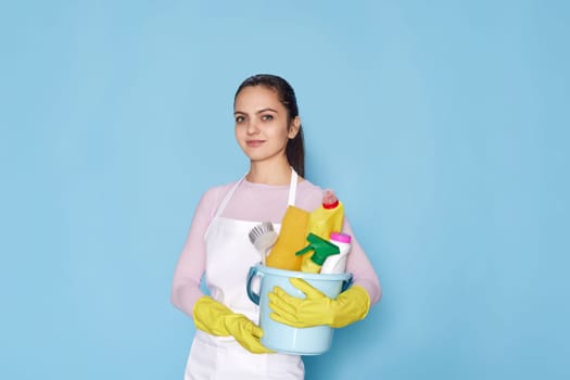 smiling caucasian woman in gloves holding bucket with cleaning supplies isolated on blue background.
