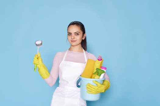 pretty woman in rubber gloves and cleaner apron holding bucket of detergents and toilet brush on blue background.