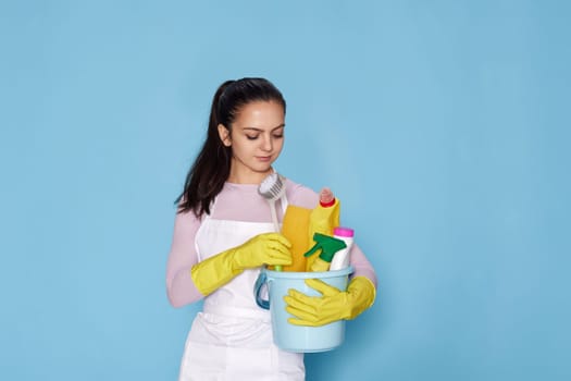 caucasian woman in gloves holding bucket with cleaning supplies isolated on blue background.
